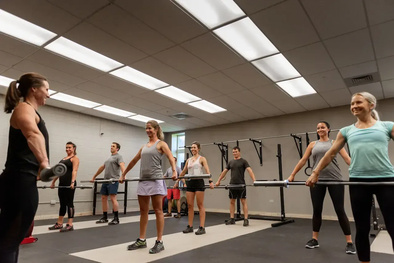 Group fitness class in a gym, led by an instructor with a barbell, students holding barbells, smiles.
