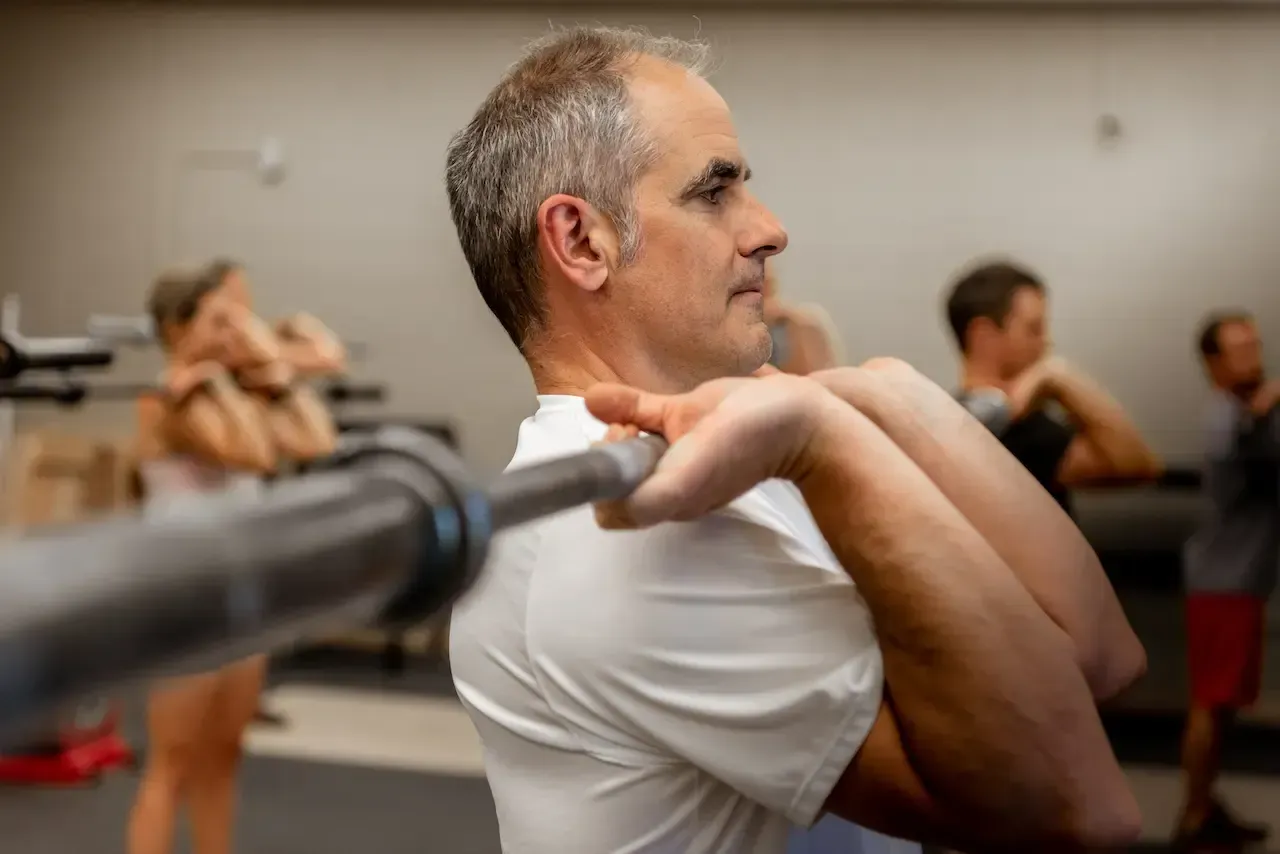 Man in white shirt holds barbell during exercise, other people in background. Gym setting.
