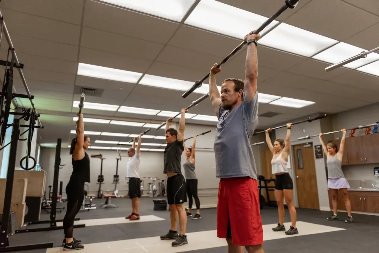 People holding barbells overhead in a gym. White walls, fluorescent lights.