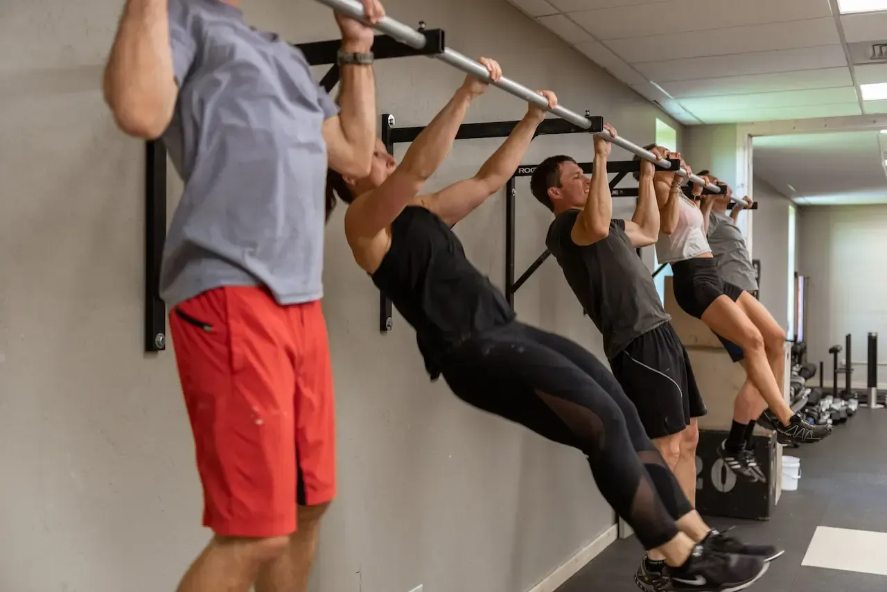 People doing pull-up exercises on bars in a gym, several are in a row, building strength and fitness.