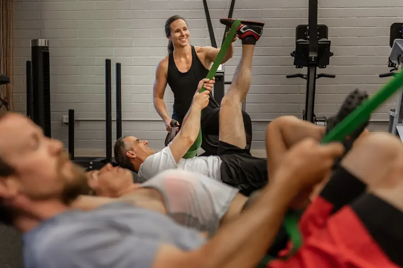A woman assists a man with a leg stretch using a green resistance band in a gym. Other men are nearby.