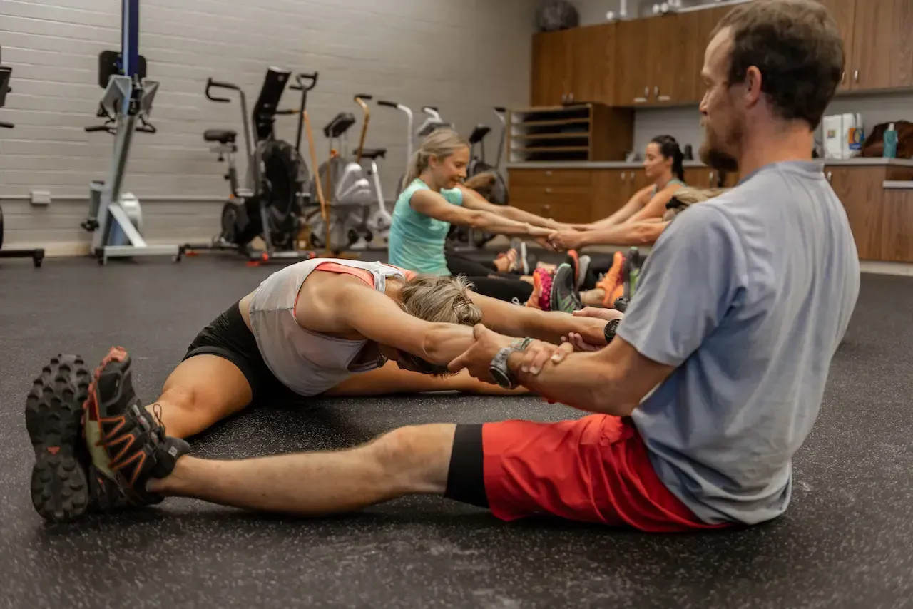 People stretching with a trainer in a gym. They are seated and holding hands while leaning forward.