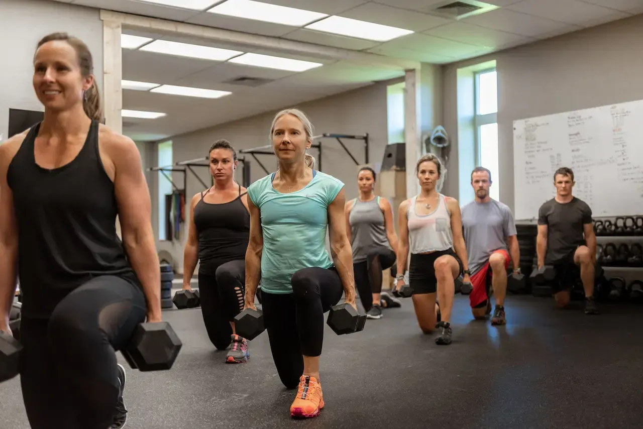 People doing dumbbell lunges in a gym. Black floor, neutral walls, natural light.