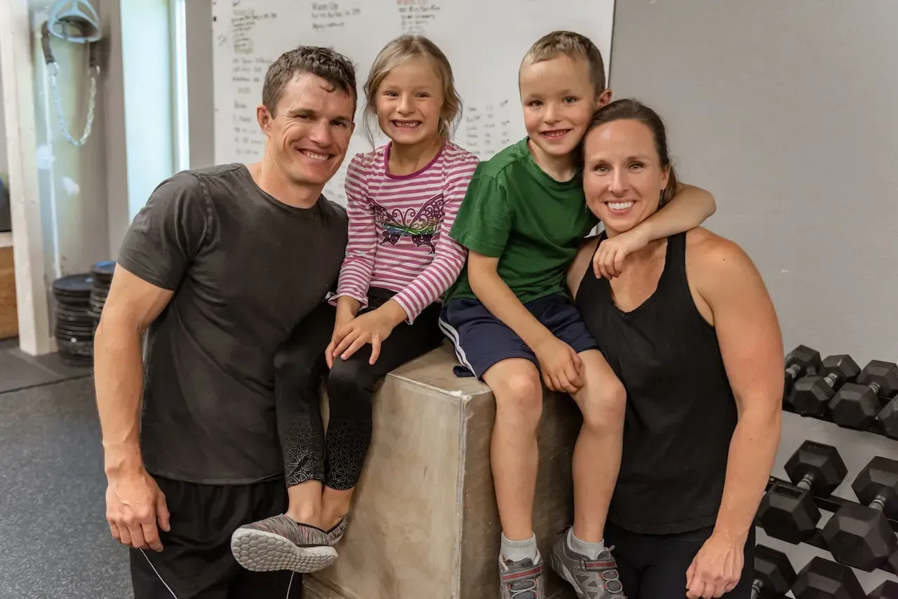 Family of four poses in a gym; two children sit on a box, adults smile with arms around them.