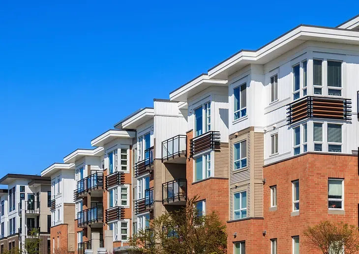 Apartment building exterior with white trim, balconies, and brick and beige siding against a blue sky.