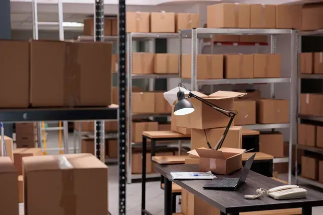 Warehouse interior with shelves of cardboard boxes, desk with laptop and lamp.