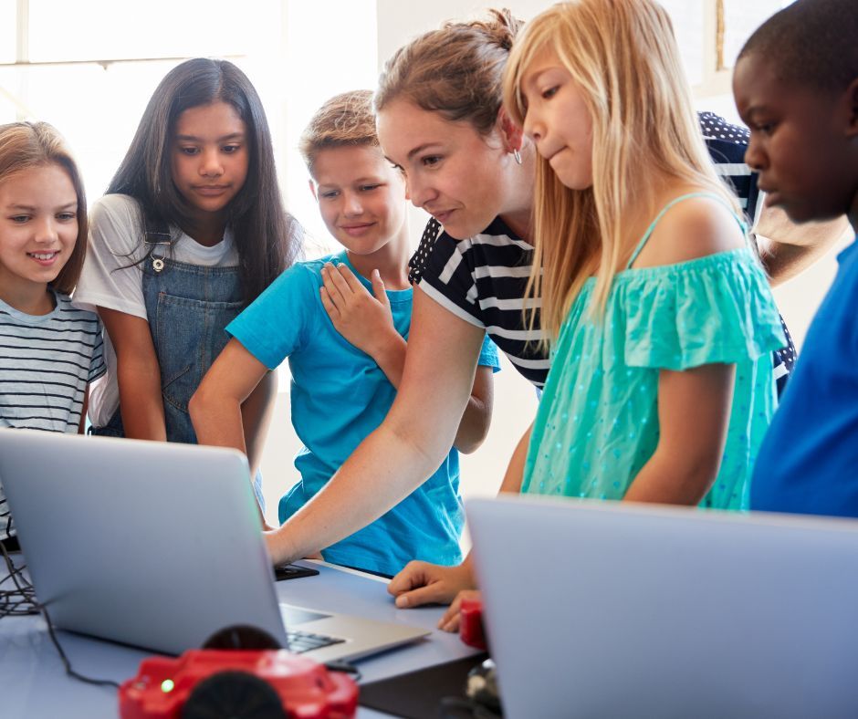 Students gather around laptops in a classroom, pointing and discussing a screen.