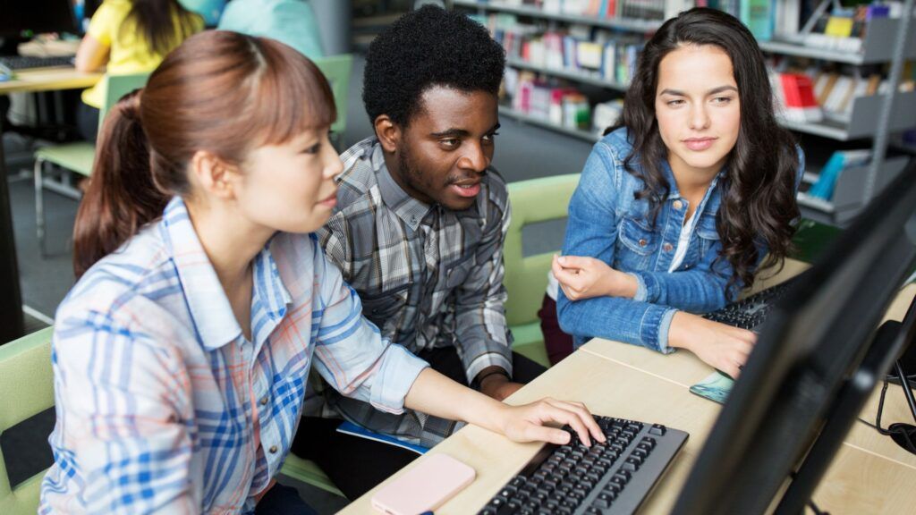 Three students collaborating at a desktop computer in a library or computer lab