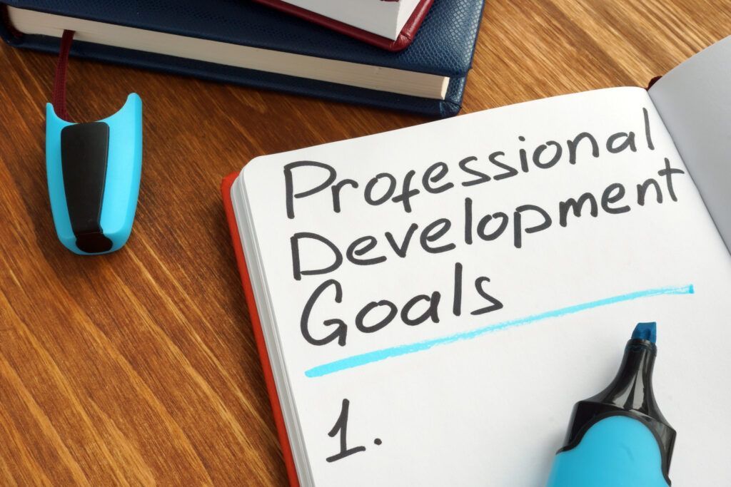 Notebook titled “Professional Development Goals” on a wooden desk with a blue pen and binder clips.