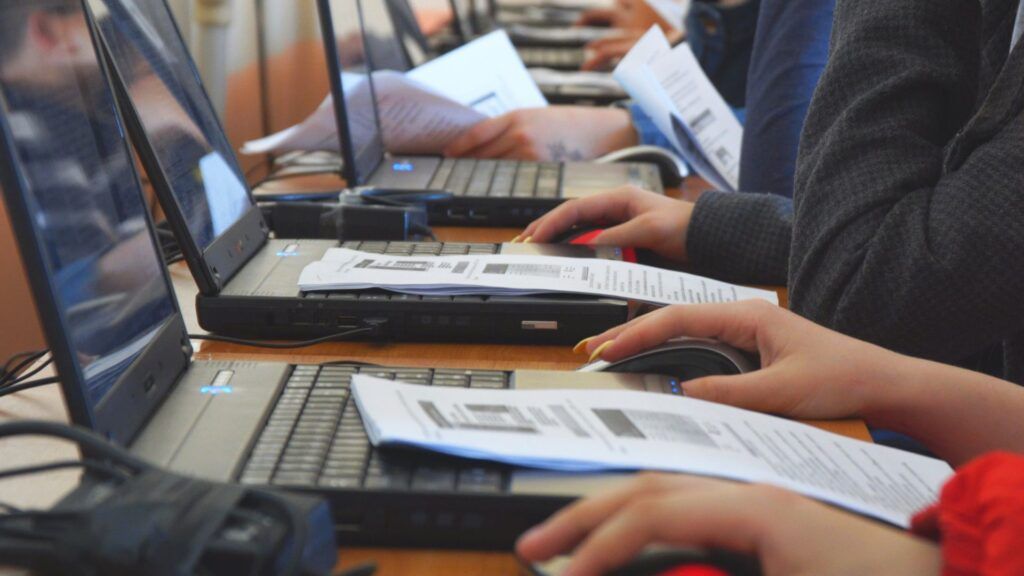 People typing on laptops at a table, with notebooks and papers in a classroom or workshop setting