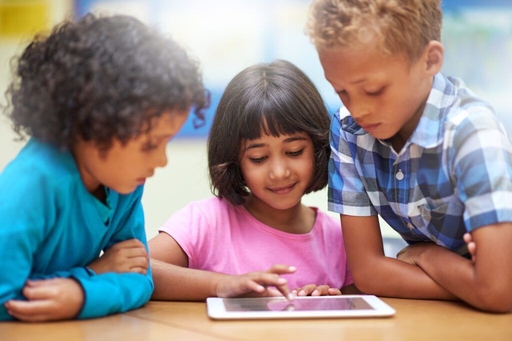 Three children lean over a tablet, smiling and interacting in a bright room