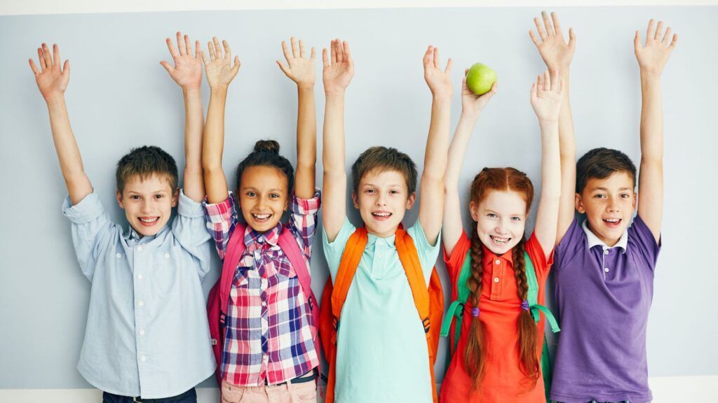 Five children standing with arms raised, smiling in a classroom, one holding a tennis ball.