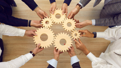 Hands from people in business attire circle wooden gears, symbolizing teamwork and collaboration.