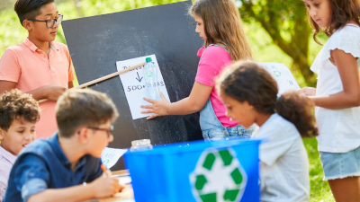 Children sorting recyclables into a blue bin outdoors at a school cleanup event