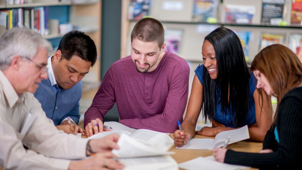 Group of people collaborating around a table, reviewing papers in a bright classroom or office setting