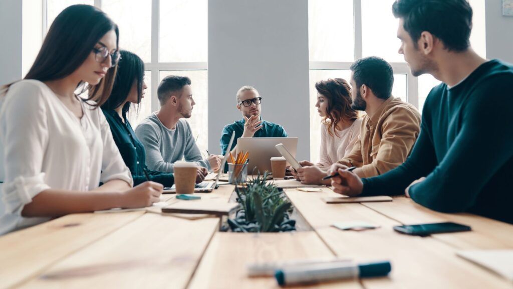 People in a meeting around a table, talking and working in a bright office.