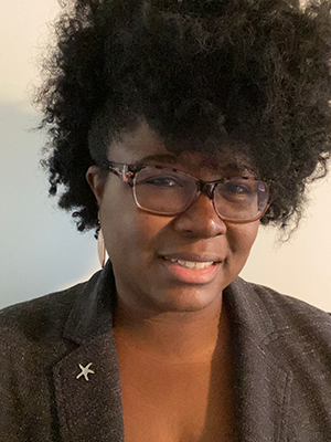 Portrait of a smiling person with glasses and curly hair, wearing a dark blazer against a light background