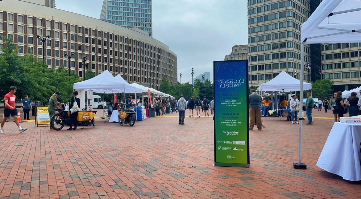 Outdoor plaza with tents, people, and a tall green sign between brick buildings and skyscrapers