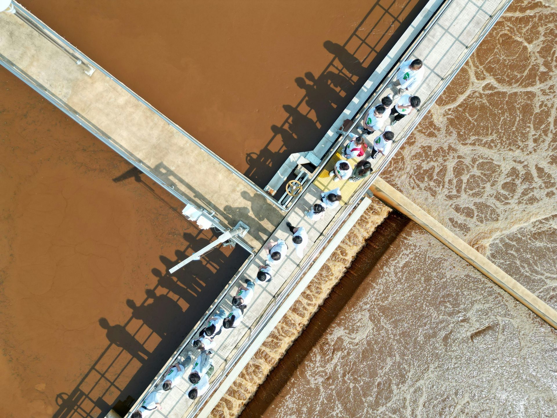 Aerial view of people walking across a diagonal bridge over brown and foamy water channels.