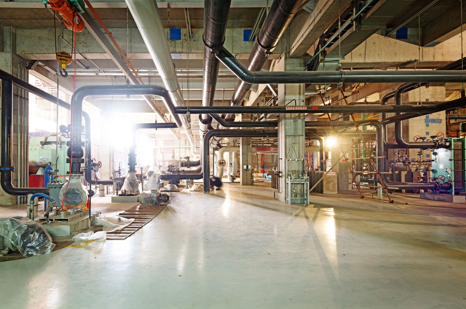 Empty industrial factory floor with large metal pipes, beams, and bright daylight streaming in from windows