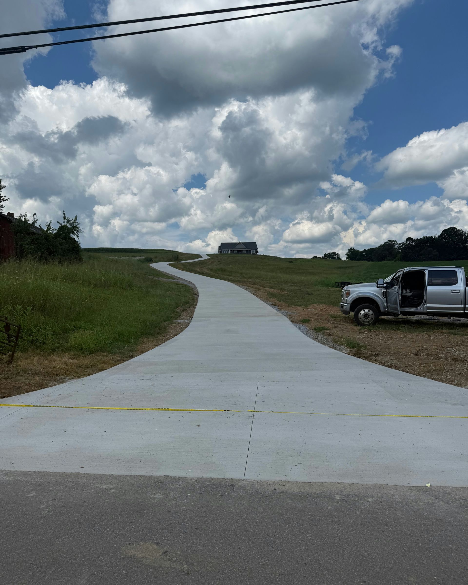 A new concrete driveway extends up a grassy hill toward a distant house, with a silver truck parked on the right.