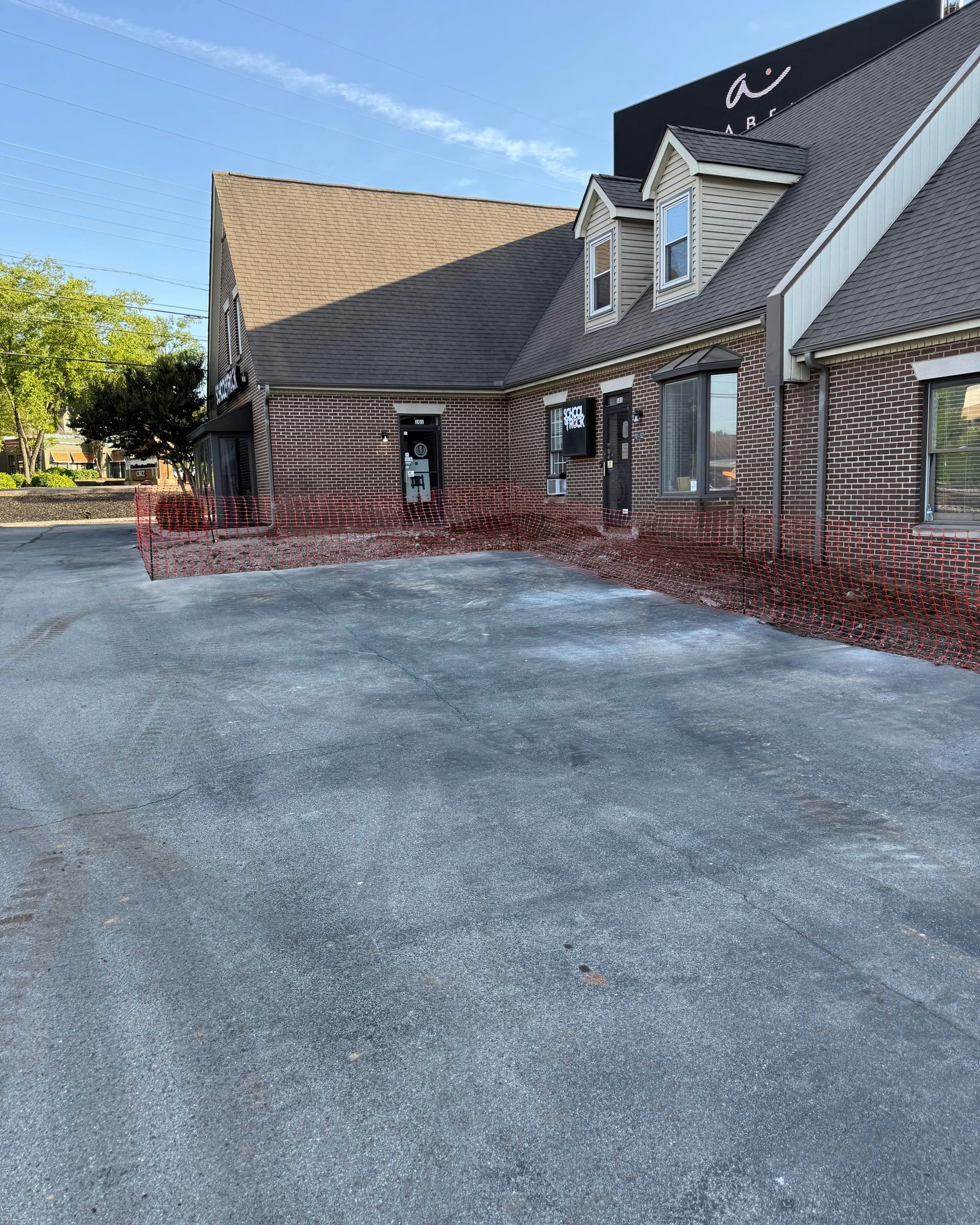 A low-angle view of a brick commercial building with a grey shingled roof under a clear blue sky, facing a paved parking lot.