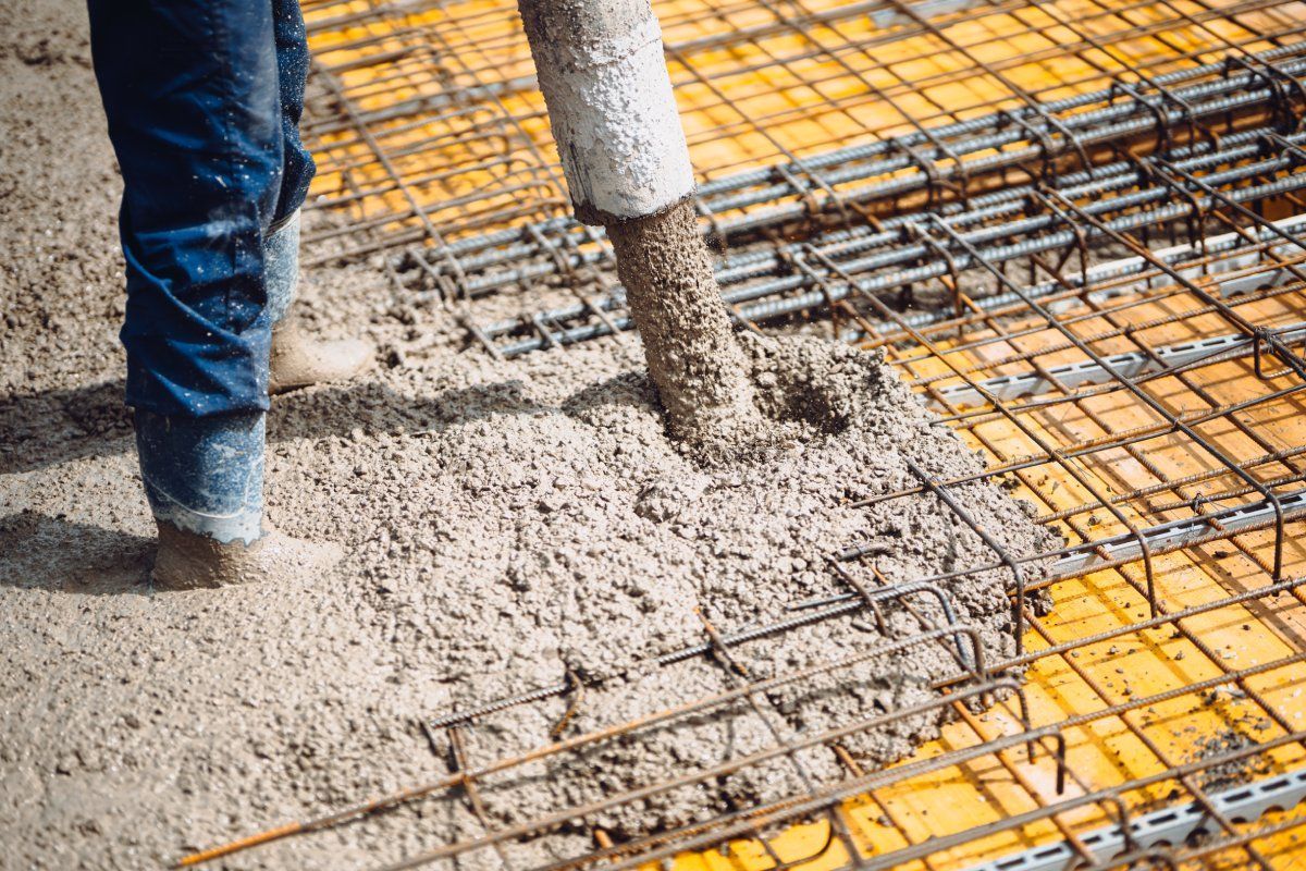 Wet concrete pours from a hose onto a yellow industrial frame reinforced with metal rebar on a construction site.