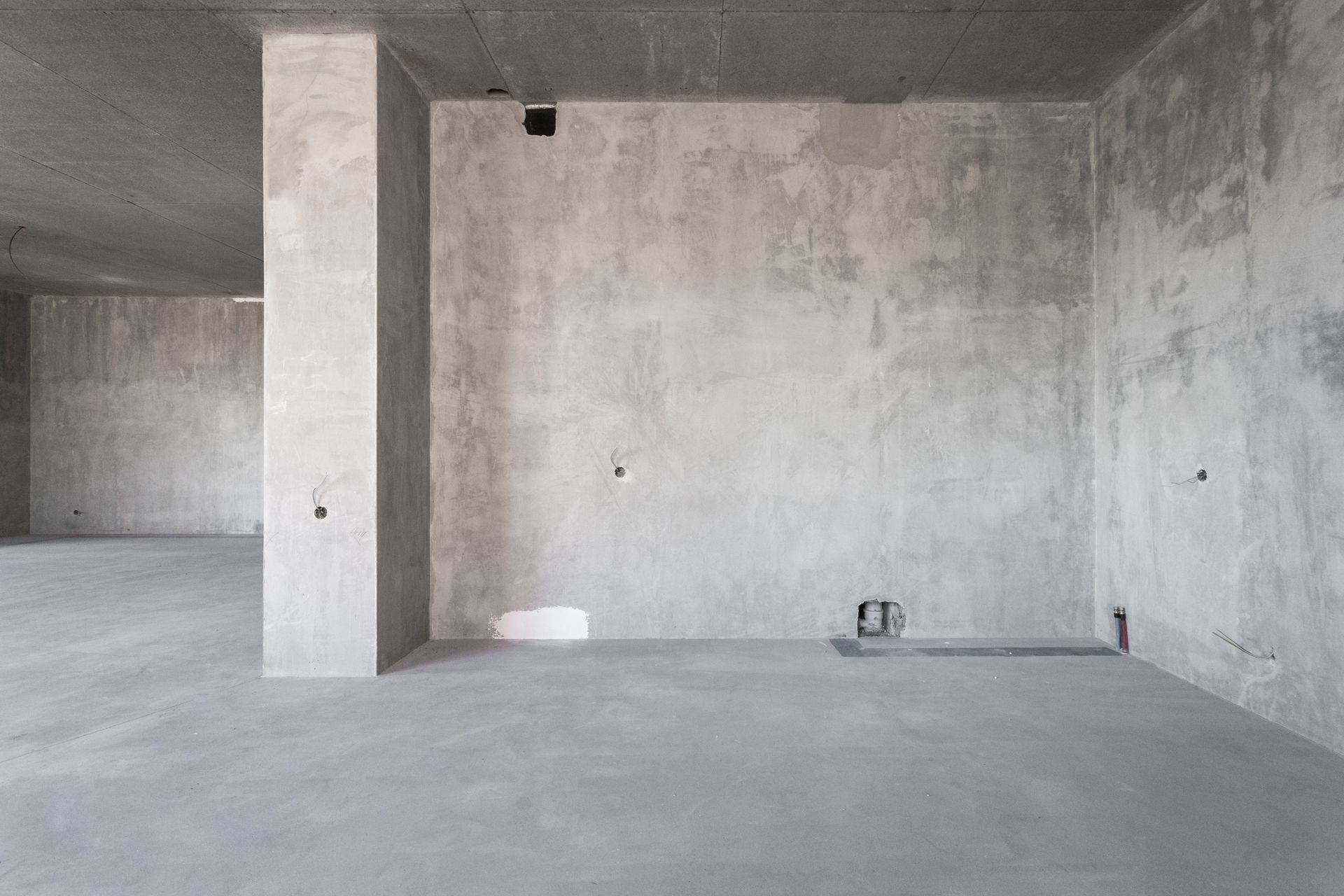 An interior view of an unfinished, minimalist room with gray concrete walls, floor, and ceiling, featuring a central pillar.