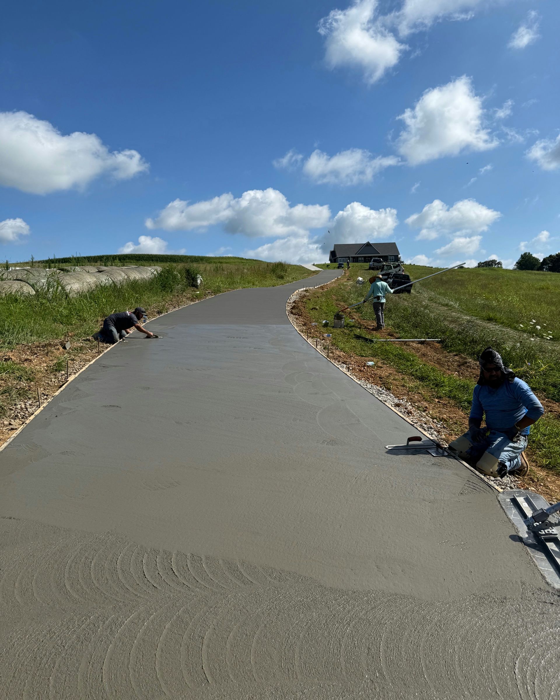 Three workers smooth a fresh concrete driveway winding up a grassy hill under a bright blue, cloudy sky.