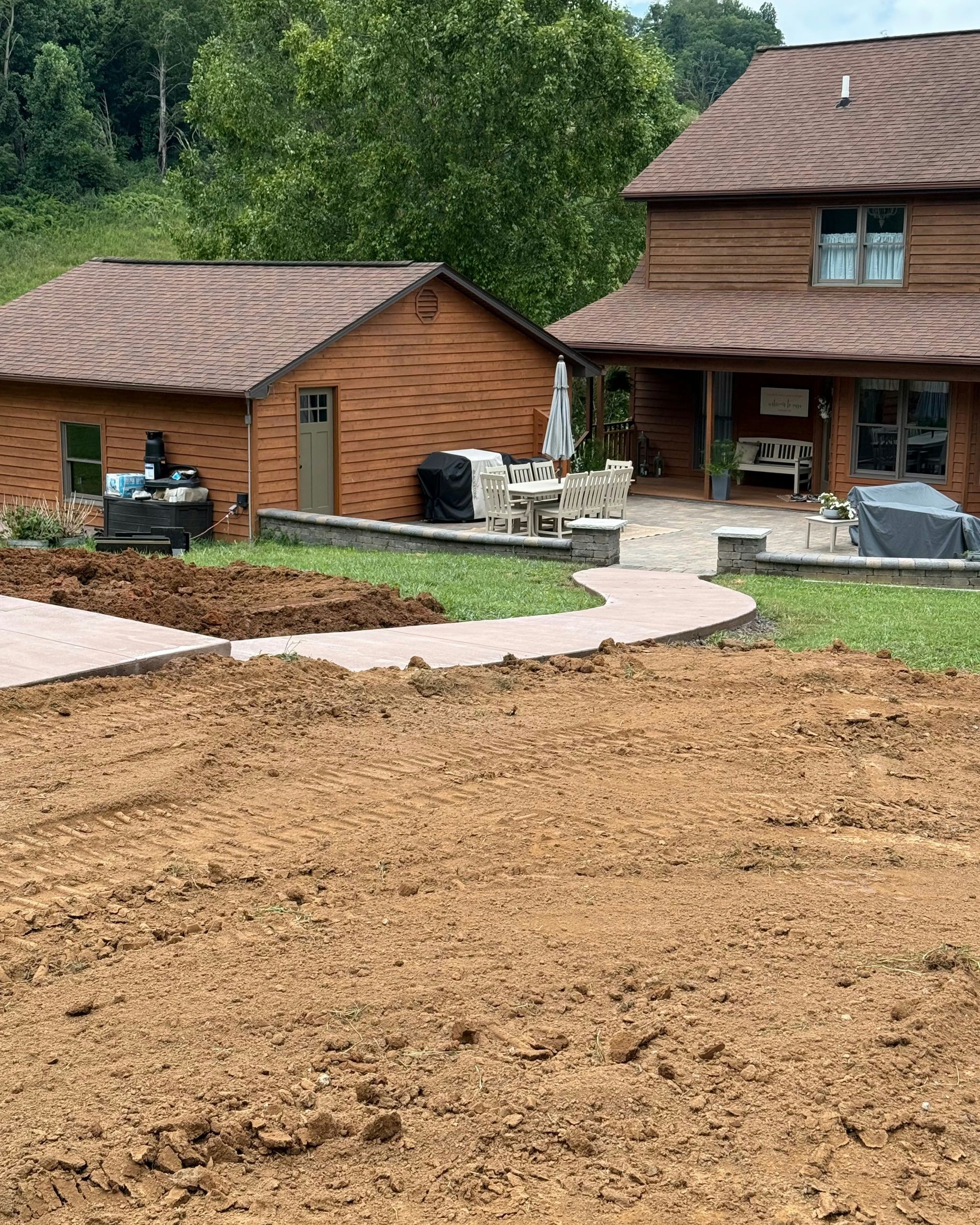 A rustic wooden house and detached garage with a patio and a large, unfinished dirt yard in the foreground.