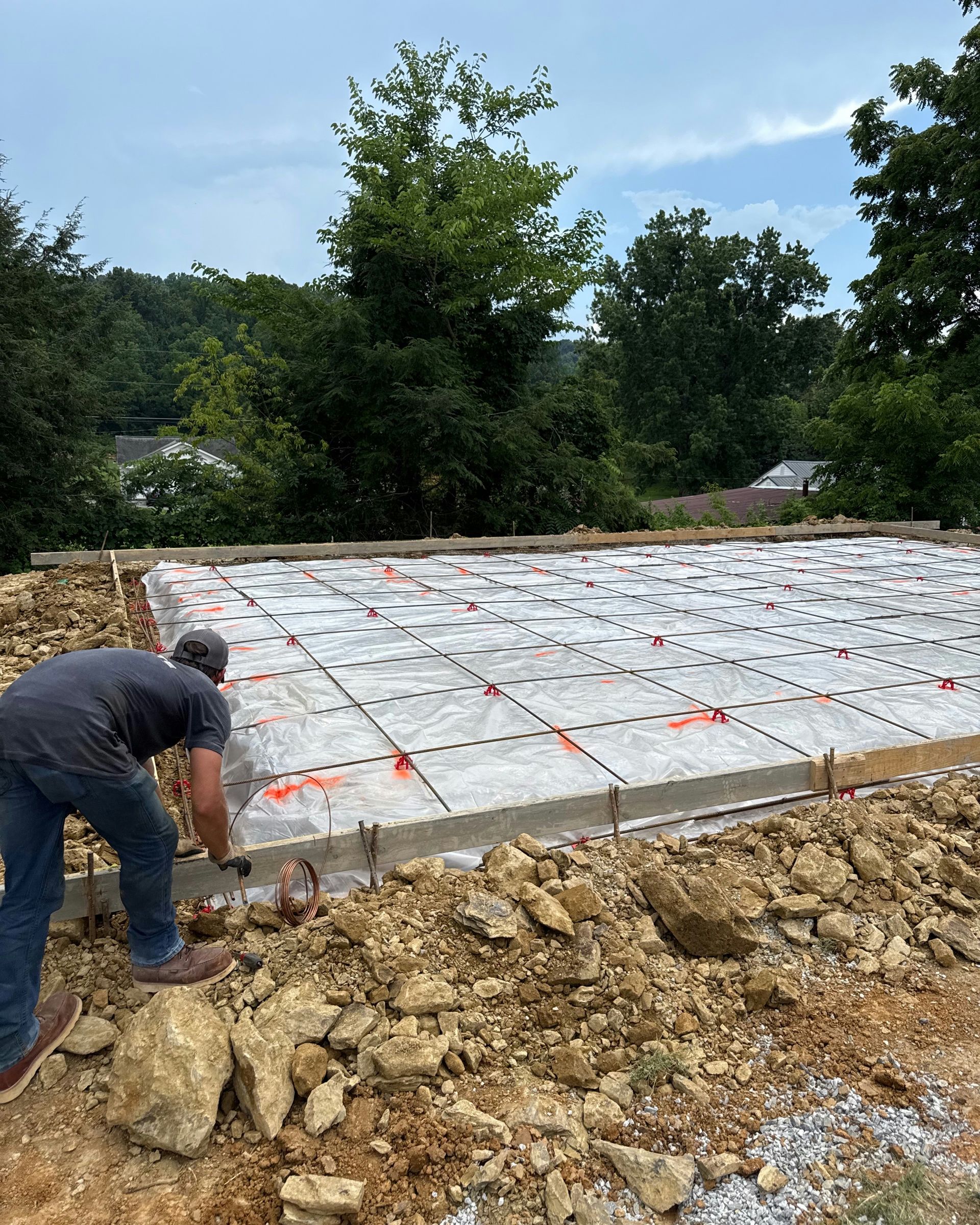 A person in a gray shirt and blue jeans works on a rectangular foundation covered in plastic sheeting at a construction site.