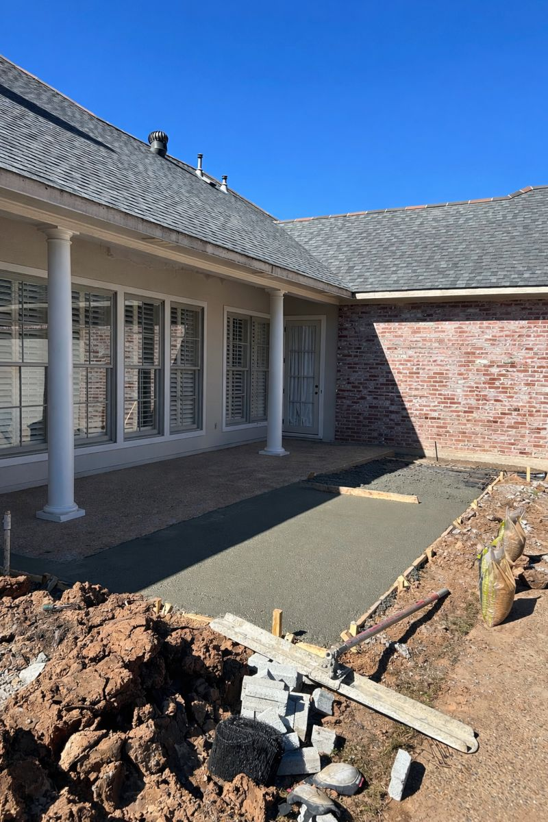 A newly poured concrete patio slab under construction next to a house with white columns and a brick exterior.