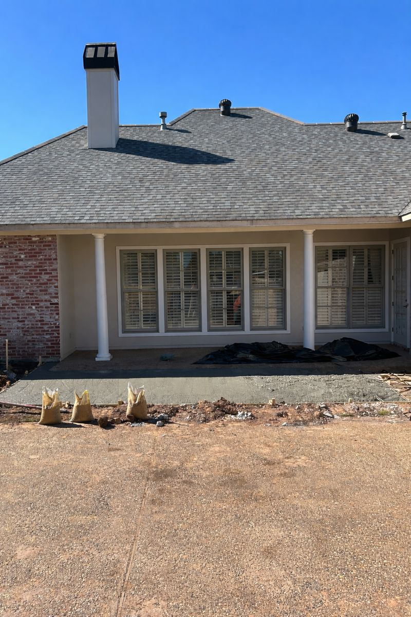 Back patio of a house with light-colored siding, a gray shingled roof, a brick corner, and large shuttered windows.
