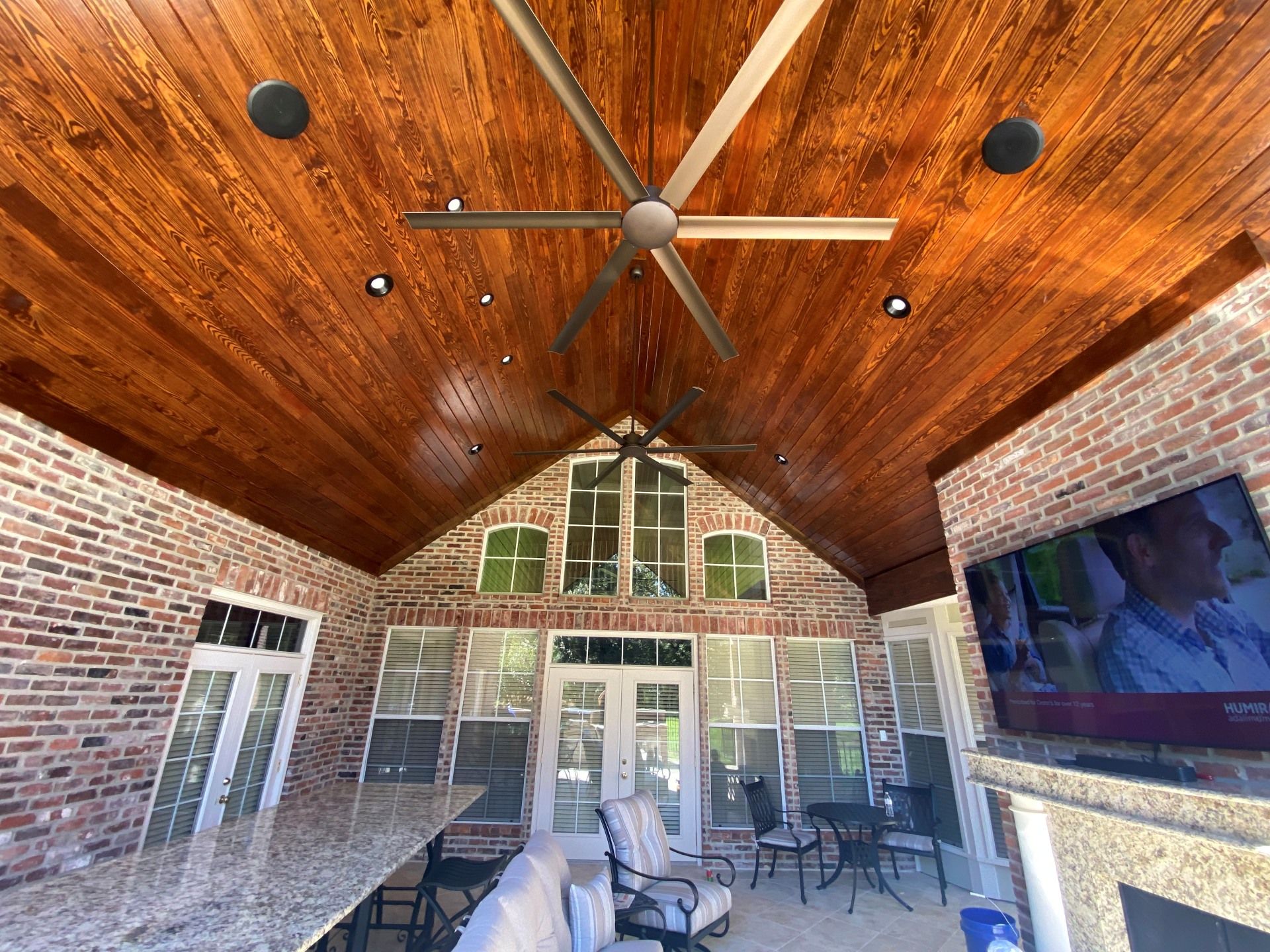A covered brick patio with a vaulted wooden ceiling, ceiling fans, recessed lighting, and an outdoor fireplace with a TV.