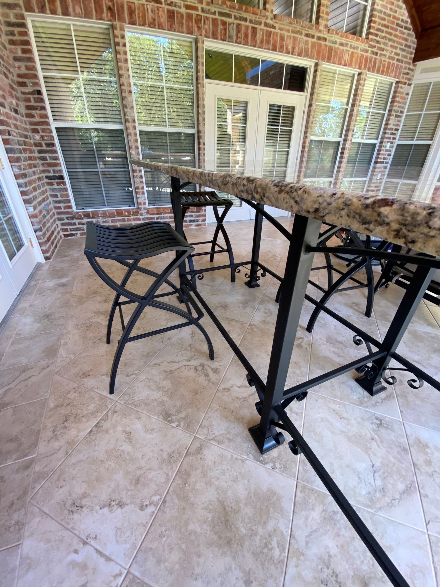 A granite-topped bar with black metal stools on a light-colored tiled patio against a brick wall.