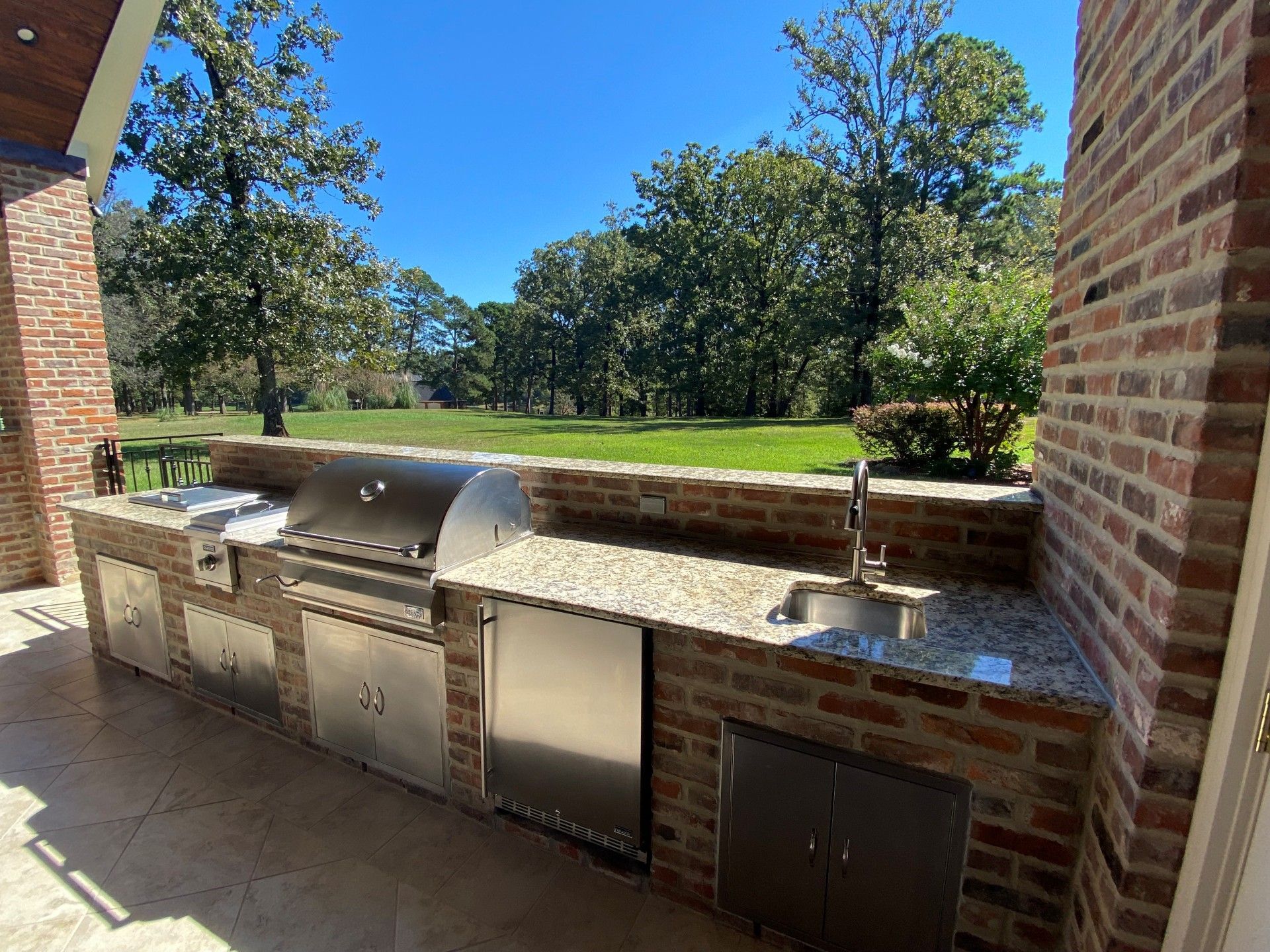 Outdoor brick kitchen featuring a stainless steel grill, counter, sink, and storage cabinets, overlooking a grassy yard.