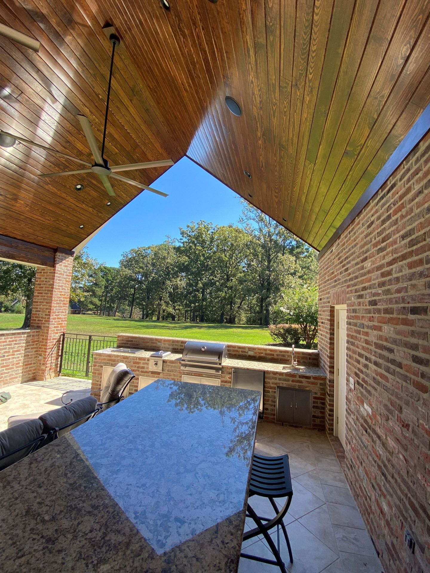 An outdoor kitchen under a wooden vaulted ceiling, featuring a granite island, brick walls, and a view of a grassy yard.
