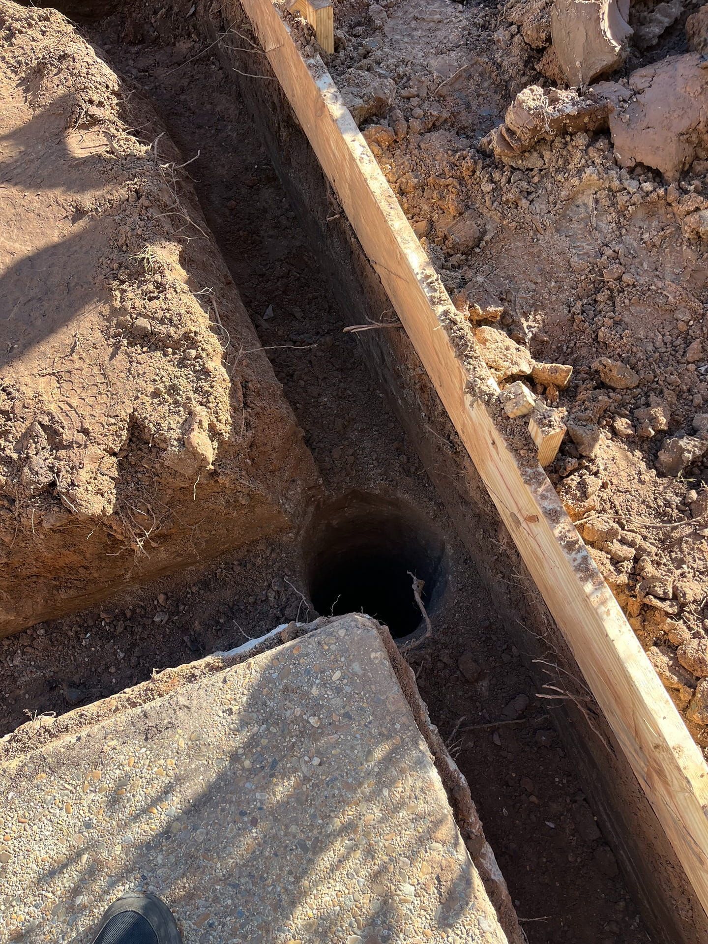 An open trench for construction dug into the dirt alongside a wooden form board and a concrete surface.
