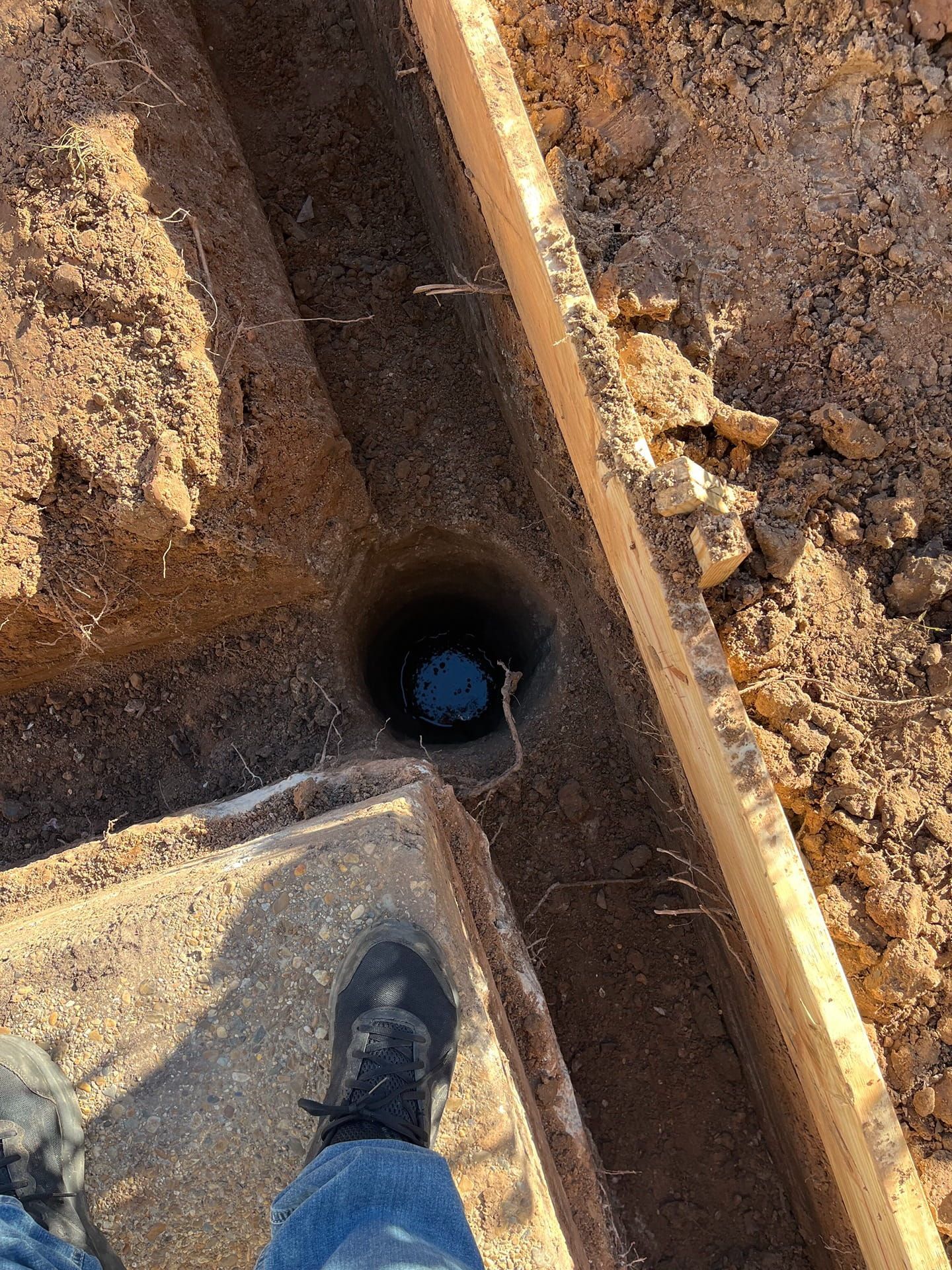 A person stands at the edge of a narrow construction trench containing a deep, water-filled hole in the dirt.