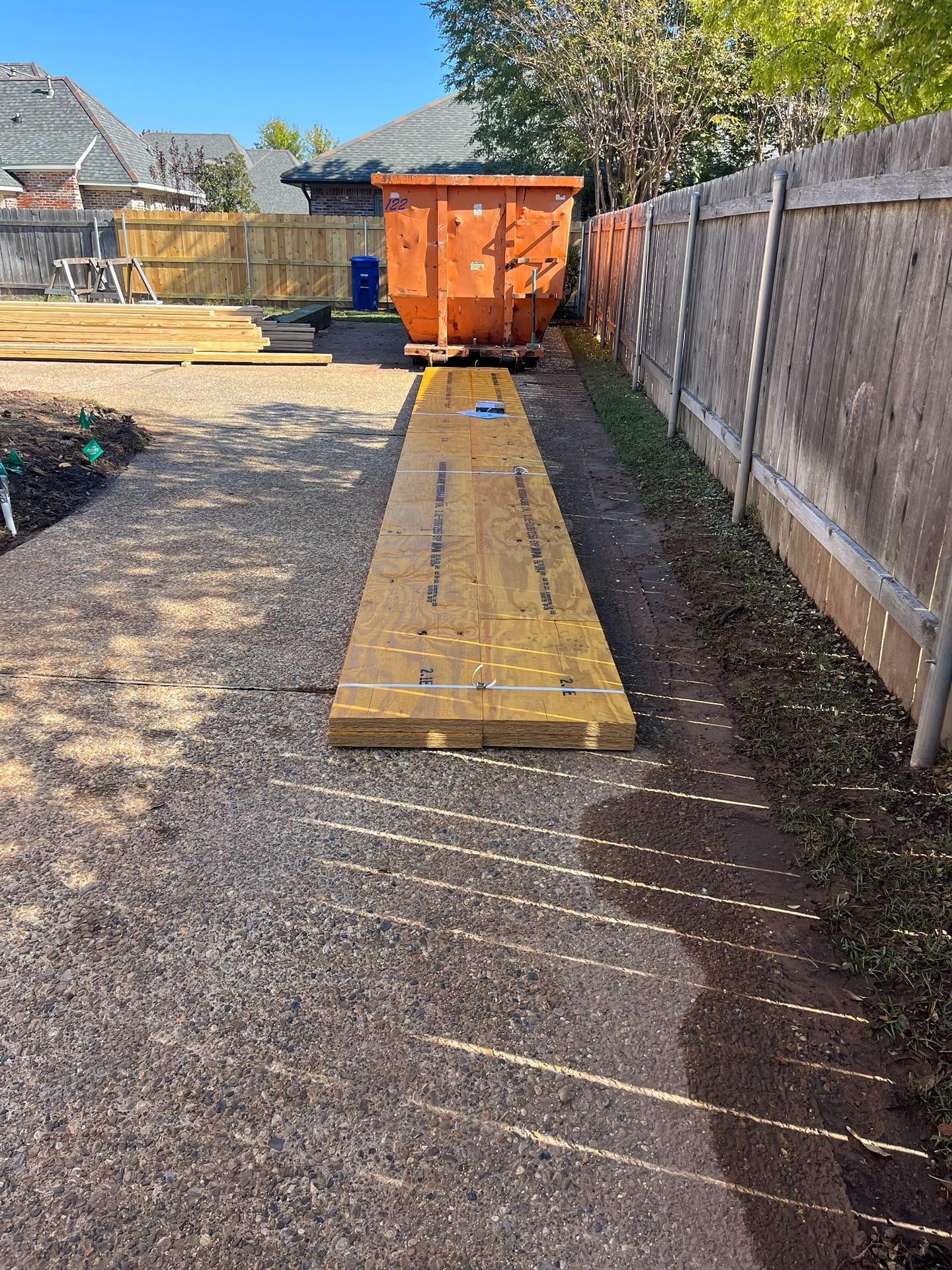 A large orange dumpster sits in a backyard next to a wooden fence, with a stack of yellow construction lumber in front.
