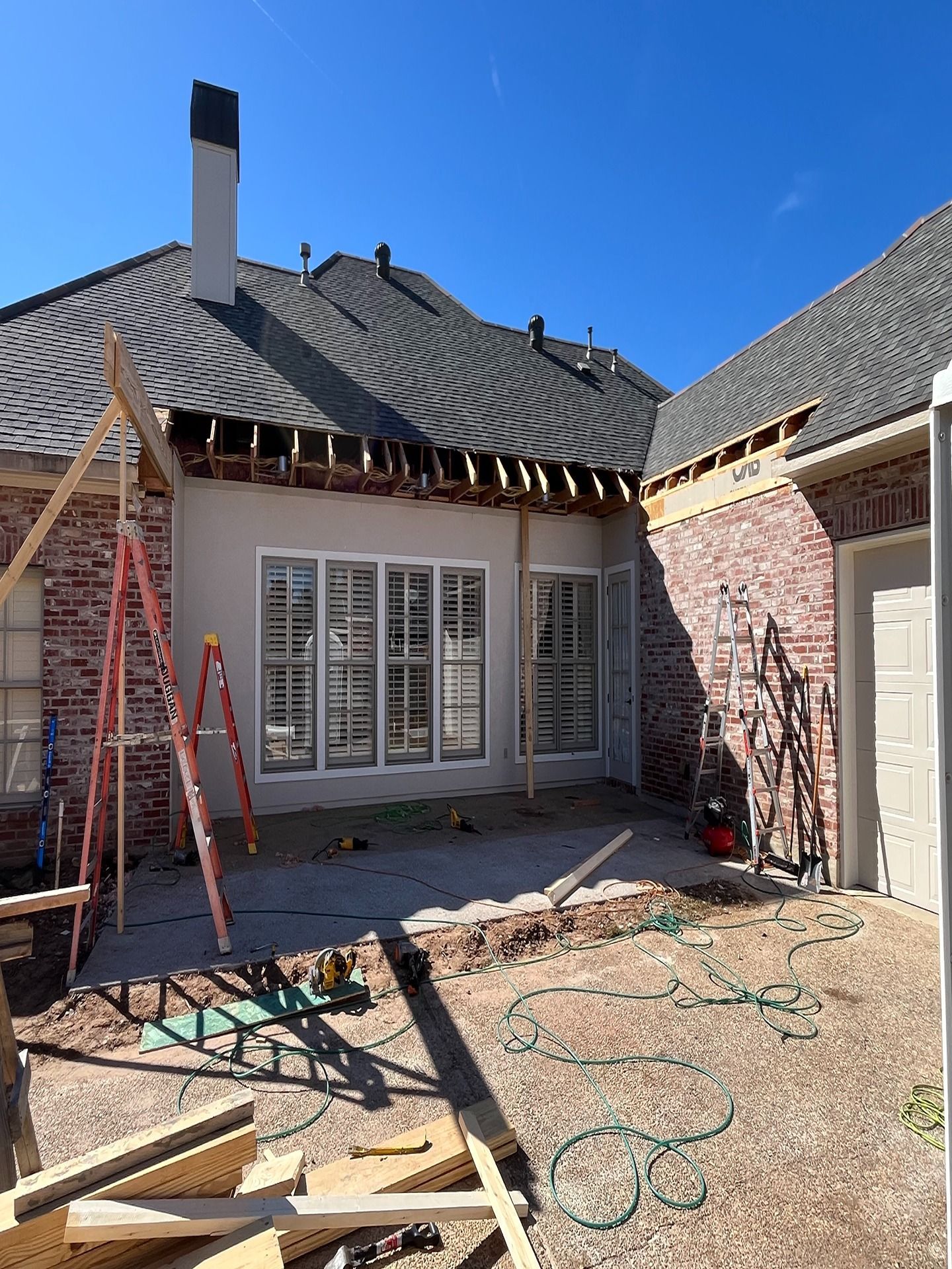 Exterior view of a home under renovation, showing exposed roof rafters above windows and brick walls, with a ladder.