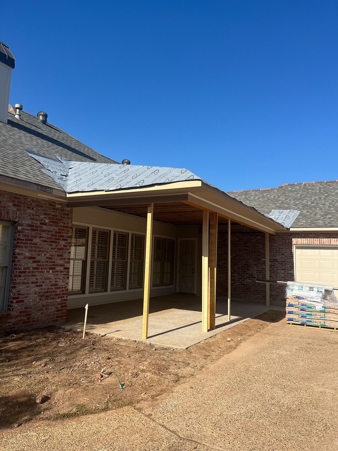 A wooden patio cover under construction attached to the back of a brick house with gray shingles under a blue sky.