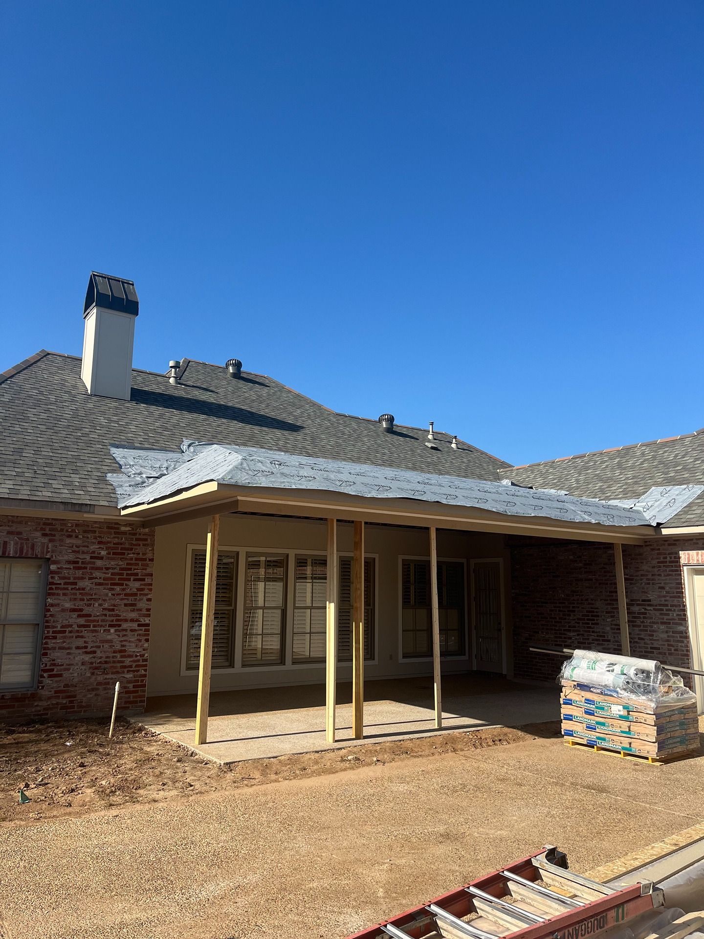 A residential patio addition under construction, featuring a wooden frame and metal roof against a brick house.