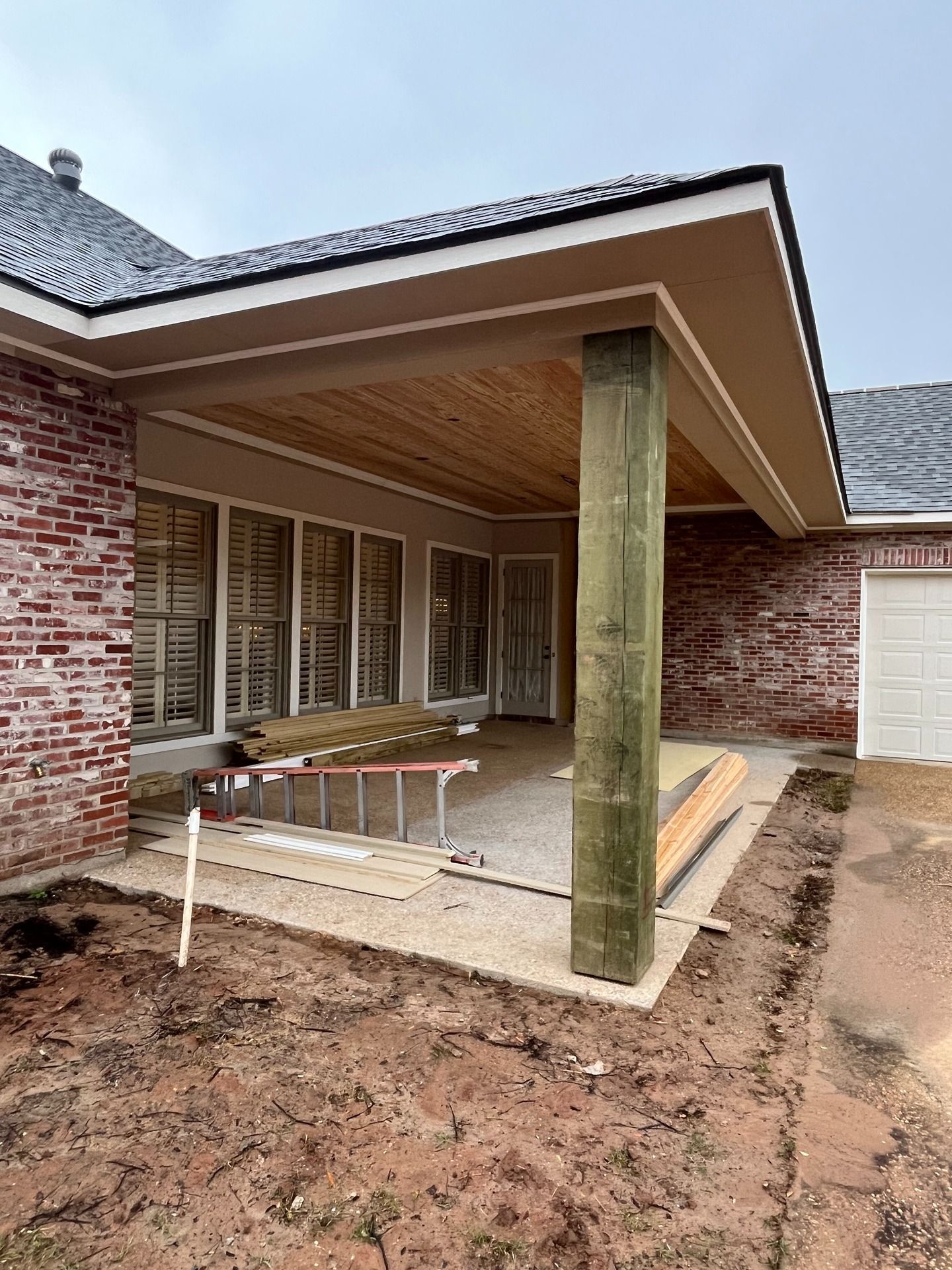 A residential back patio under construction with brick exterior, a wooden support post, and a slatted wood ceiling.