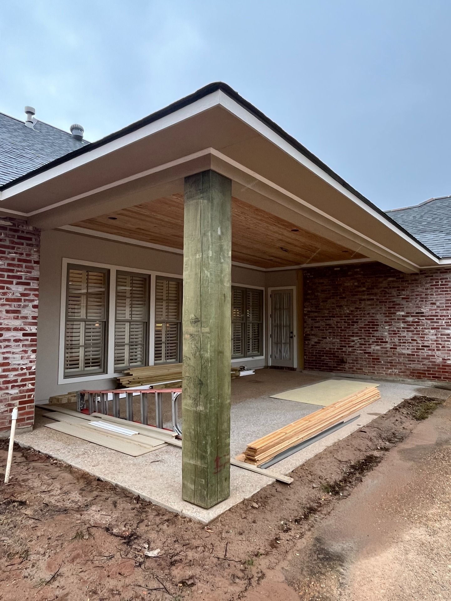 A covered patio under construction with a large wooden support post, brick walls, and exposed ceiling rafters.