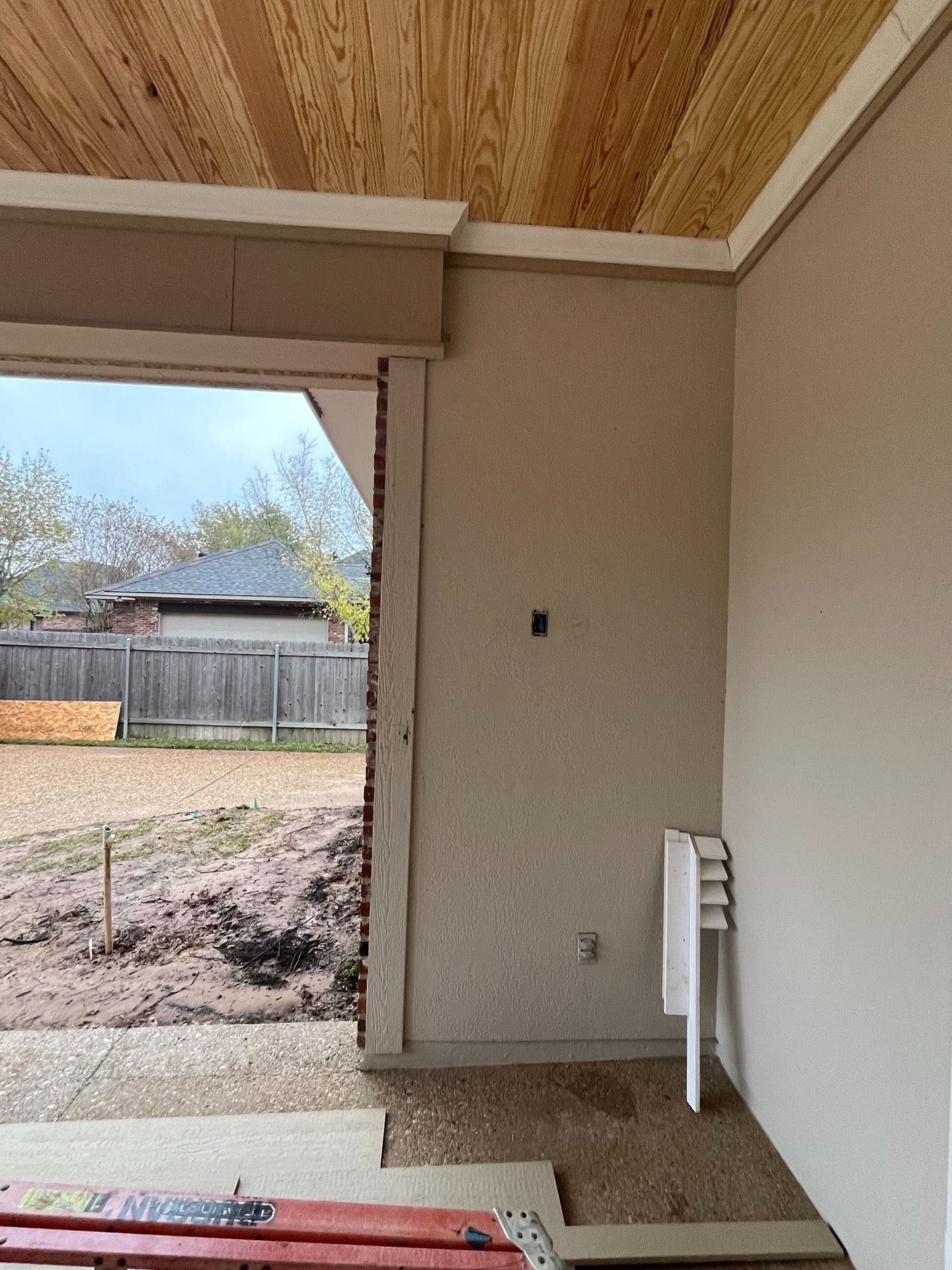 An unfinished indoor area featuring beige walls, a wood-paneled ceiling, and a doorway opening to a backyard.