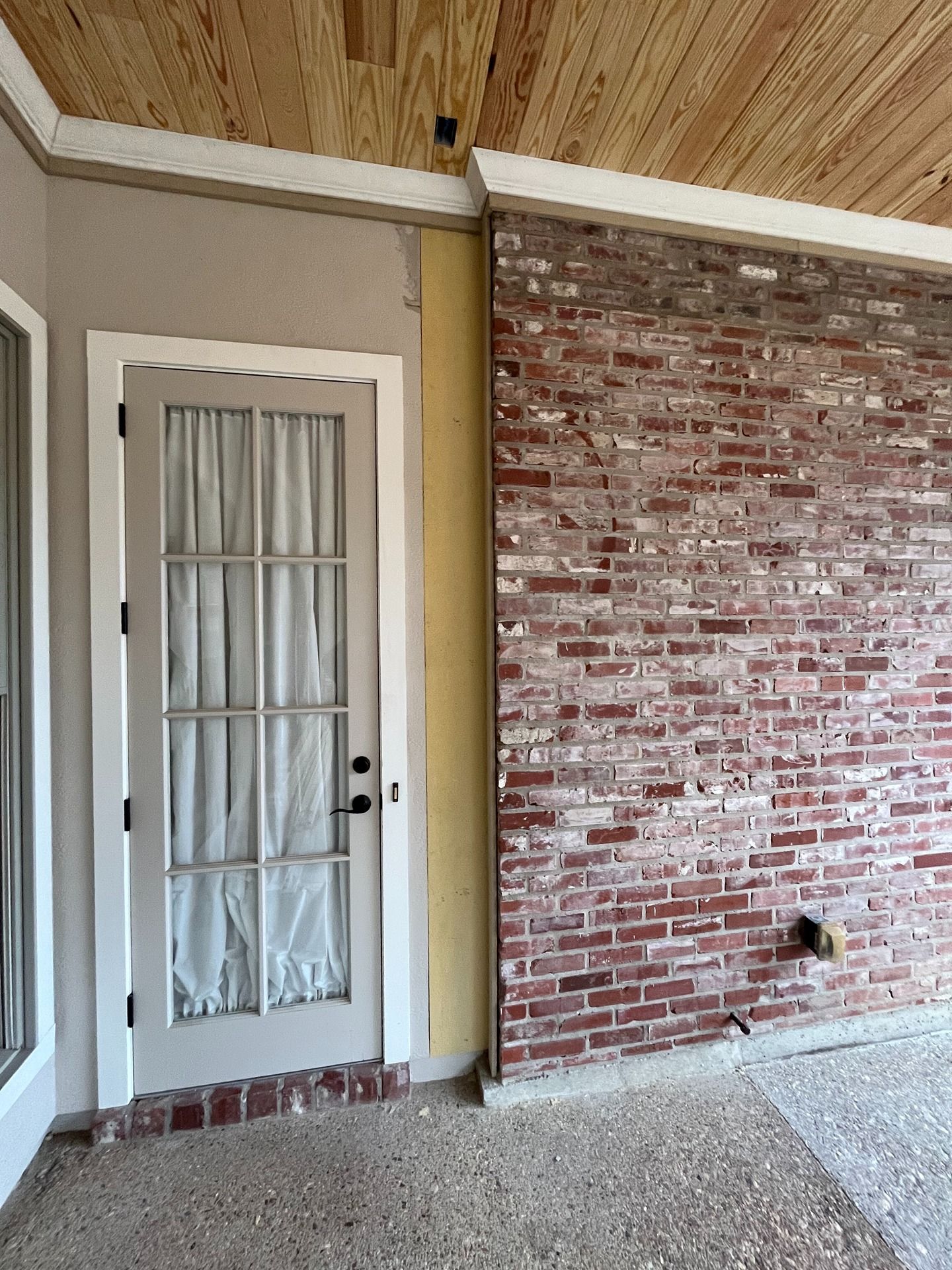 A glass-paneled door with white trim beside an exposed red brick wall on a concrete porch under a wood-paneled ceiling.