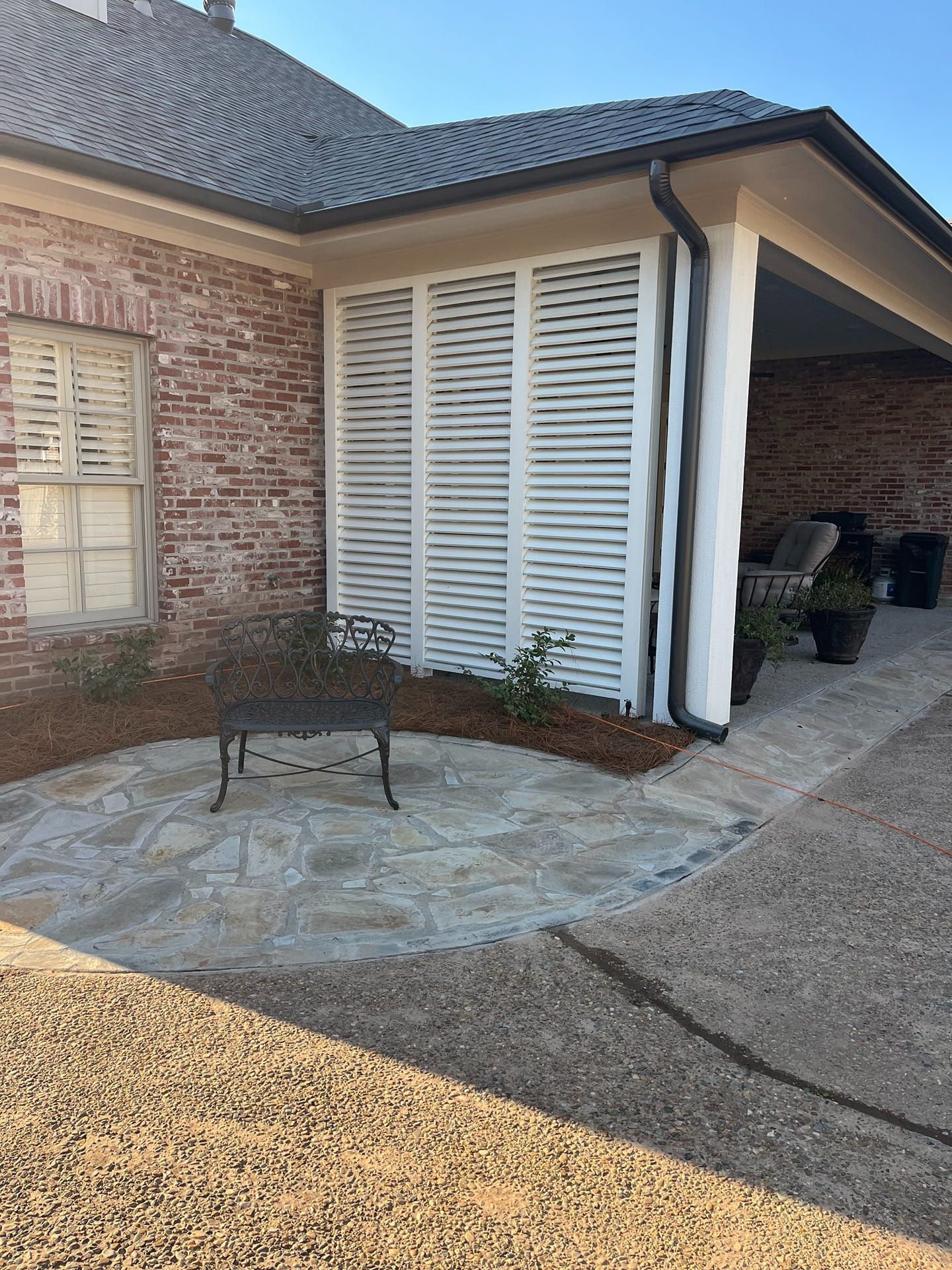 A brick home with a white shutter screen, stone patio, and a metal bench in the yard.