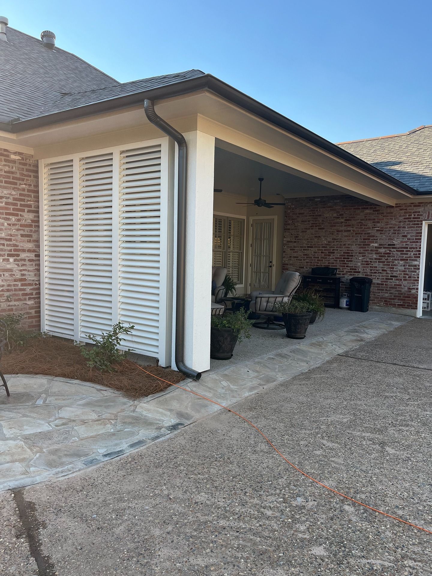 A brick house features a covered patio with white louvered privacy screens, outdoor seating, and a stone floor.
