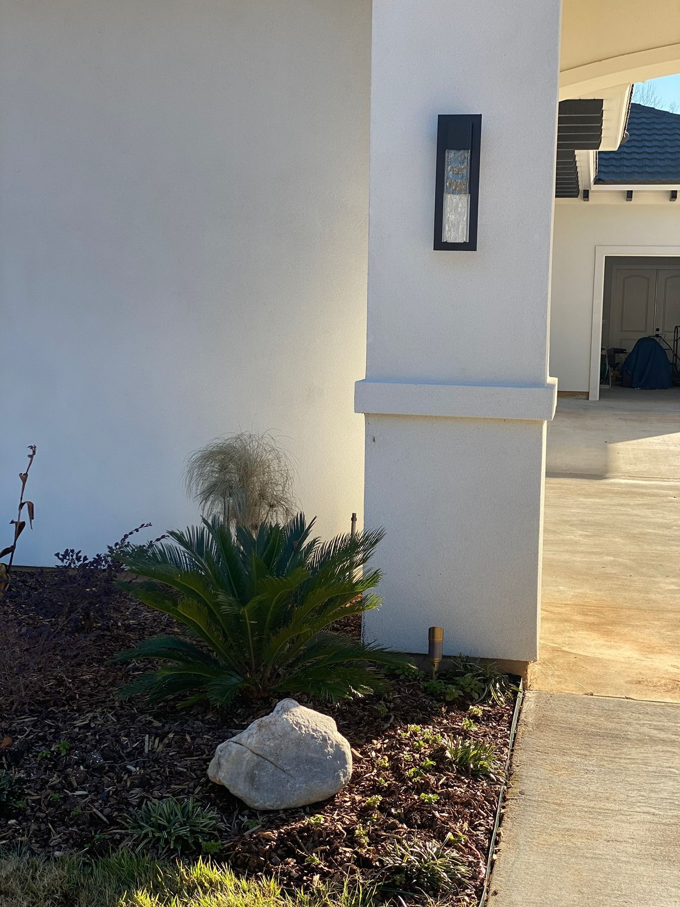 A white exterior wall featuring a modern wall lantern, a low-growing cycad plant, and a large stone in the garden bed.